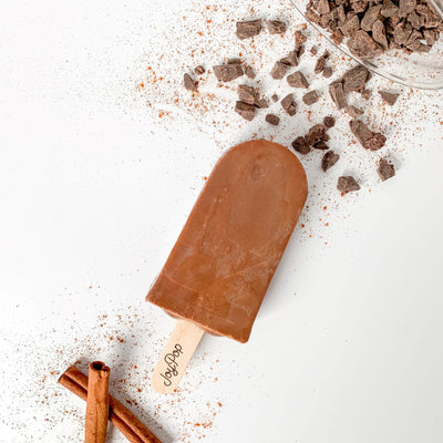 Mexican hot chocolate pop from The Joy Pop Co next to cinnamon sticks and cinnamon spice and a glass jar of chocolate chunks that is spilling out on a white background