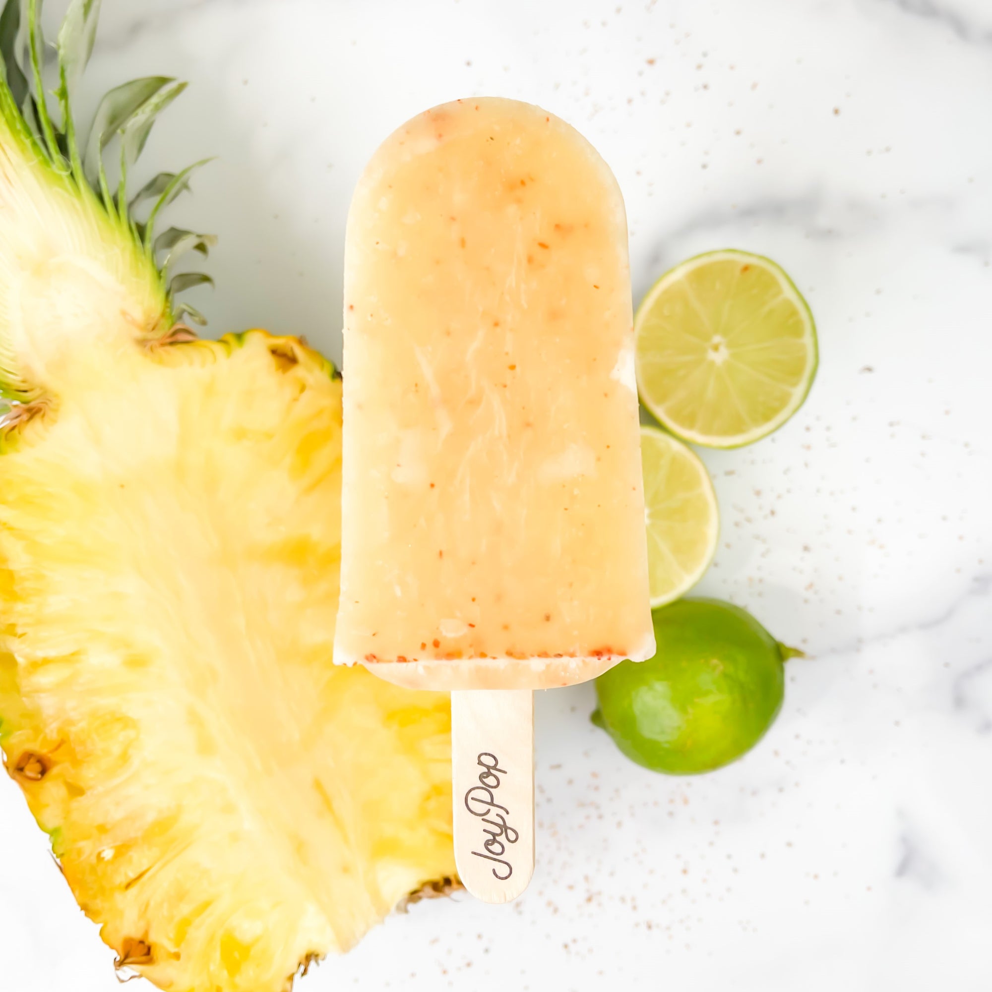Pineapple Chili Lime Frozen Pop by The Joy Pop Co next to a whole lime and cut limes and a half pineapple and chili spices scattered on a white marble background