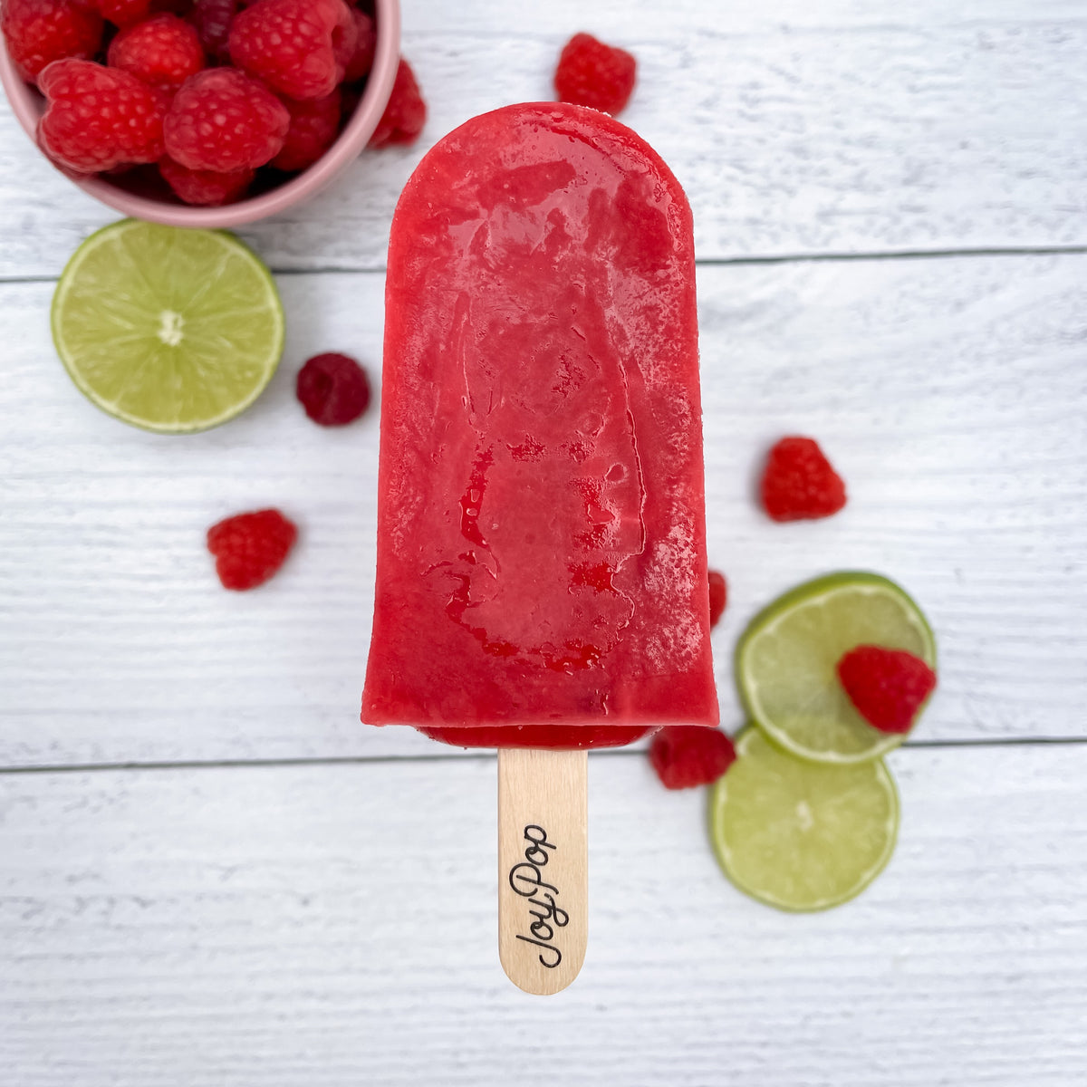 Red raspberry joy pop with a bowl of raspberries and slices of lime on a white wood background