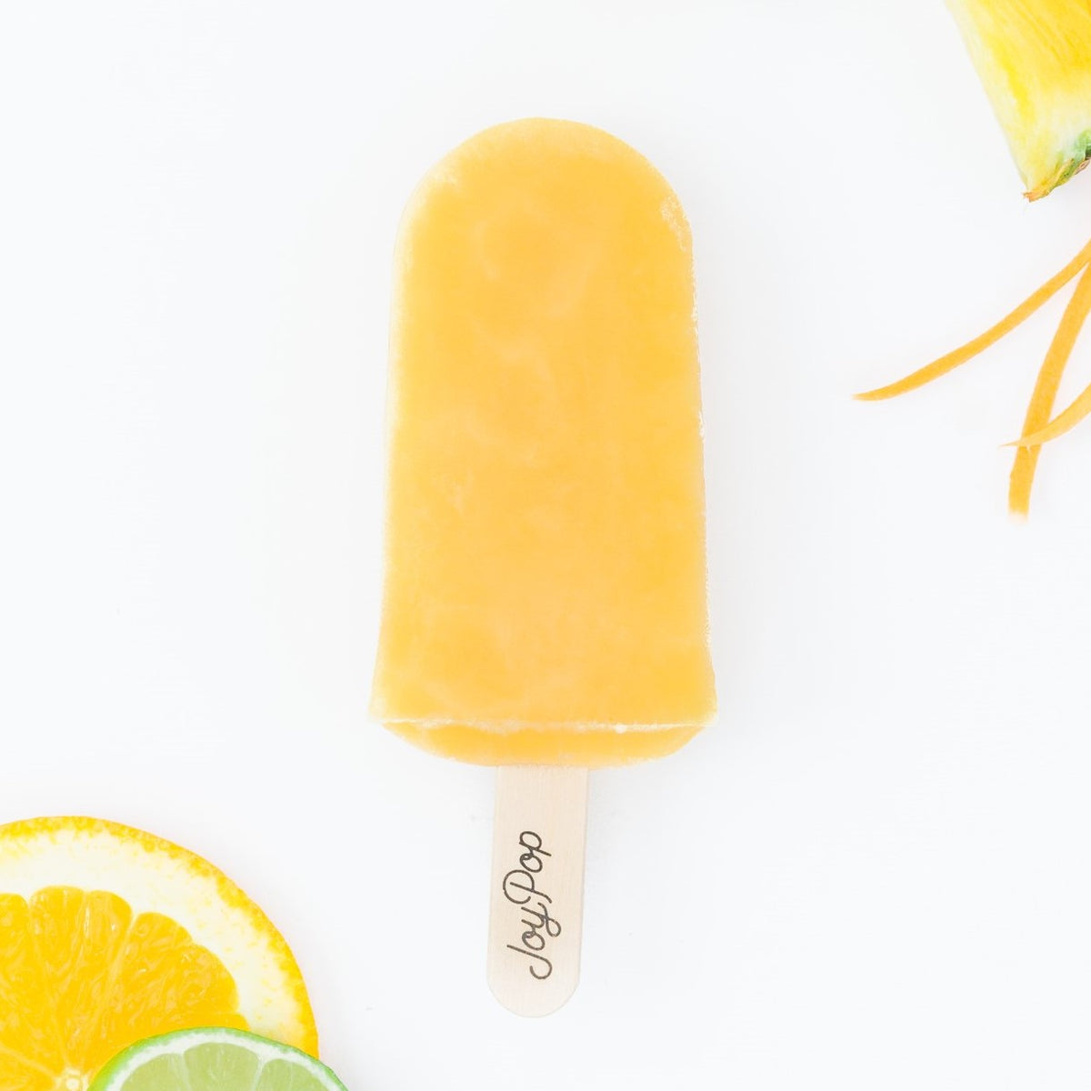 Wellness Blend frozen pop by The JoyPop Co with sliced pineapples fresh coconut a lime slice and carrot slices and a sliced orange on a white background