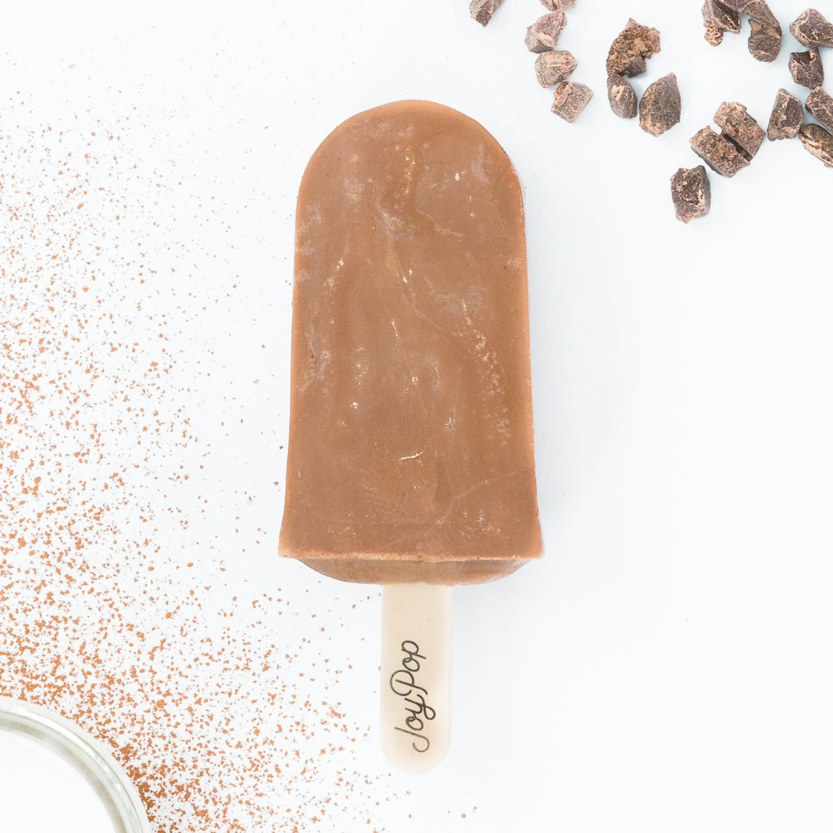 Chocolate Fudge Joy pop ice pop in the middle with chocolate pieces at the top right and a cup of cream inside a dusting of cocoa powder in the bottom left