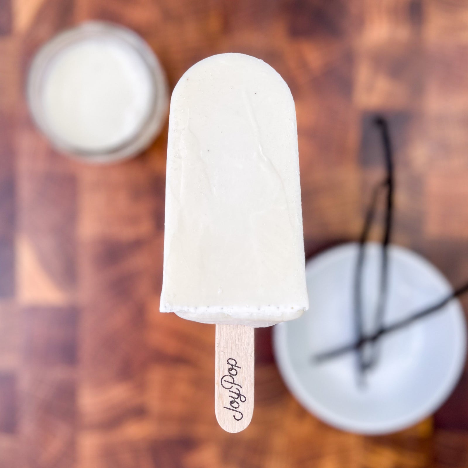 Vanilla Bean pop from The Joy Pop Co with a glass of cream and a bowl of vanilla beans in the background on a dark walnut checkered cutting board