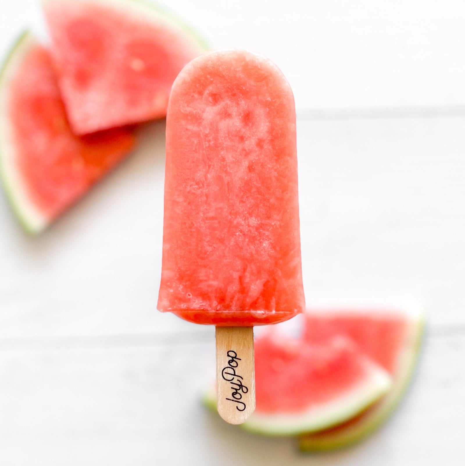 Watermelon frozen pop by The JoyPop Co next to lime slices and watermelon slices on a white background